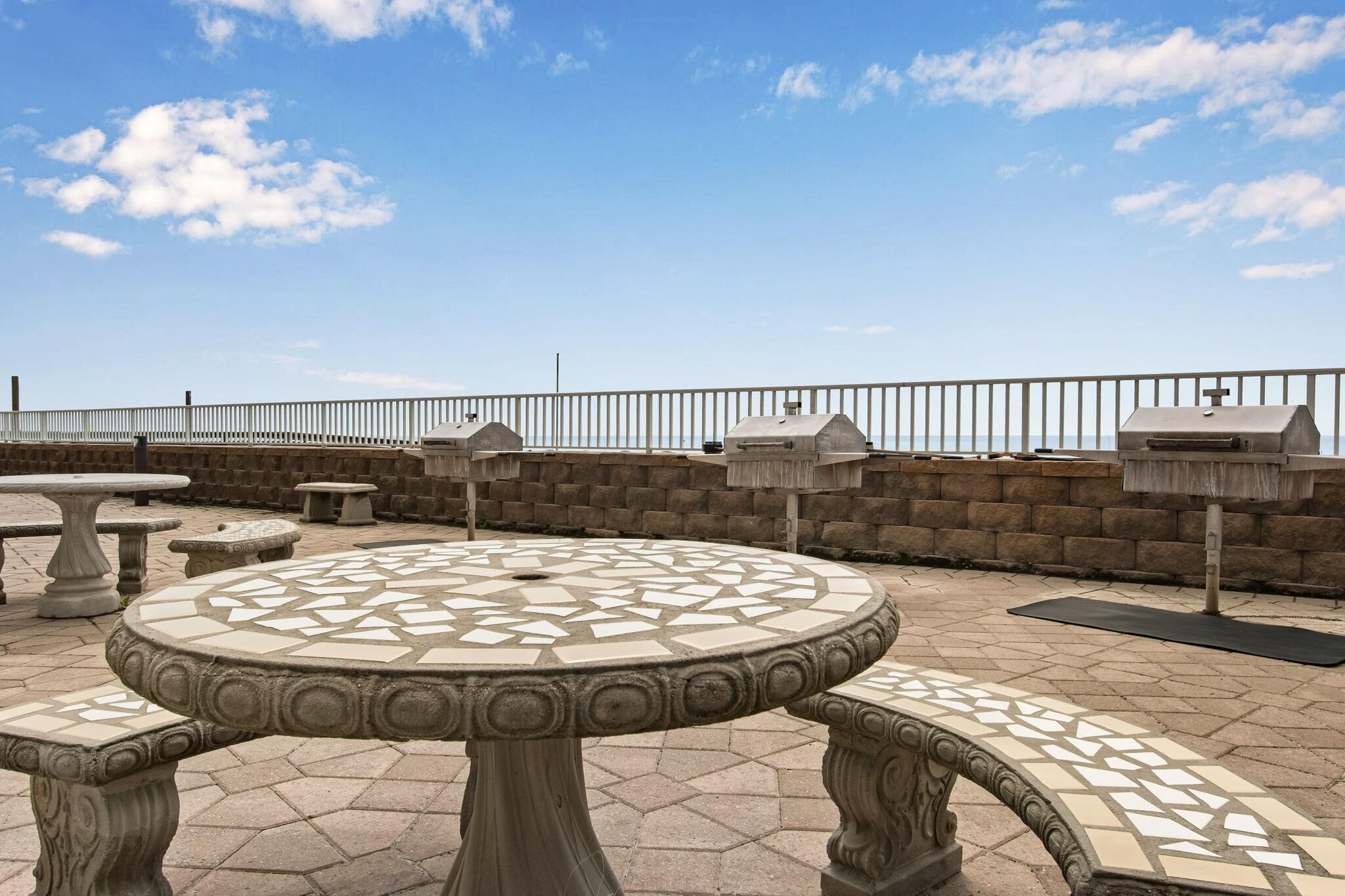 A rooftop patio with stone tables and benches, a low stone wall, and a metal railing.
