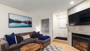 A modern living room with a sectional sofa, a coffee table, a fireplace, a mounted TV, and a large landscape painting. - Private Hot Tub | Pool | Free Parking | Sauna (Whistler)
