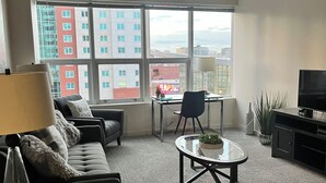 Un salon moderne avec un canapé, une table basse, un téléviseur et une vue sur le paysage urbain grâce à de grandes fenêtres.
