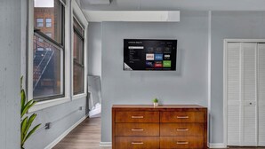 A modern living room with a mounted TV displaying streaming services, a wooden cabinet, and a large window with a view of a building. - Cozy studio in delightful Cleveland with gym, parking and AC (Cleveland)