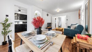 A modern kitchen with white cabinets, a microwave, and a sink. There is a dining table set with plates, cutlery, and a vase with red flowers. A blue sofa and a bookshelf are in the living area.