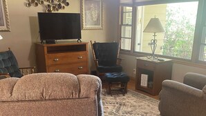 A living room with a sofa, armchair, and ottoman, a television on a wooden cabinet, a patterned rug, and a window with a view of greenery. - Cozy, spacious house located near historic downtown near shopping and dining. (Watertown)