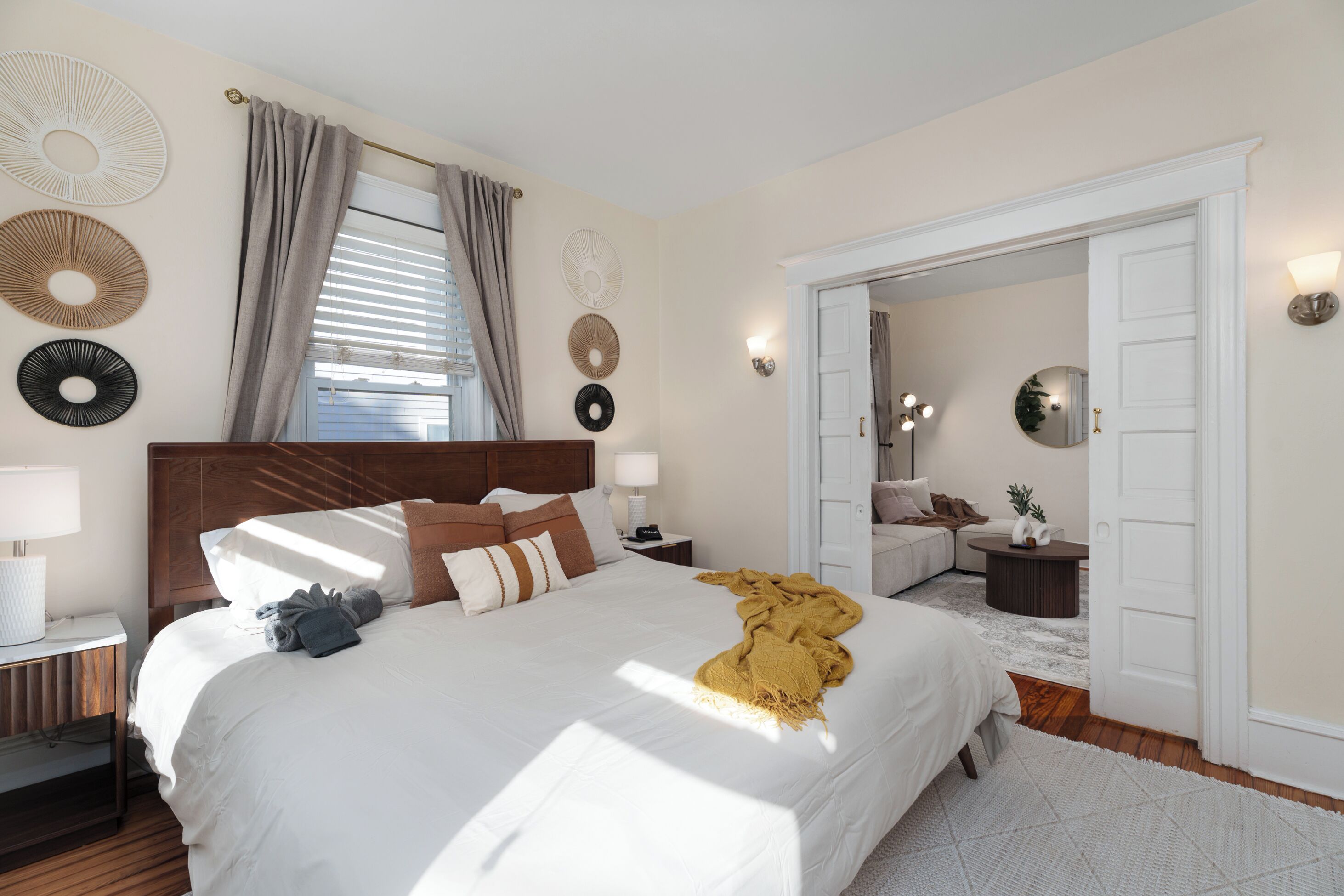 Un dormitorio bien iluminado con una cama grande, mesitas de noche, un tocador, un espejo y una puerta que da a otra habitación.