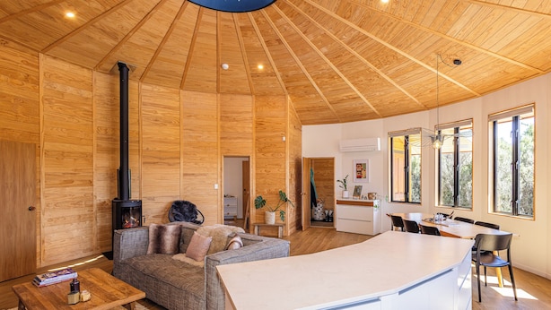 Ein geräumiger Wohnbereich mit hoher Holzbalkendecke, Kamin, einer Couch, einem Couchtisch und einer Küchenzeile mit großem Fenster.