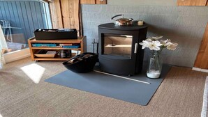Ein gemütliches Zimmer mit Holzbalkendecke, einem schwarzen Herd, einem Holzregal mit Gegenständen, einer Matte auf dem Boden und einer Vase mit Blumen.