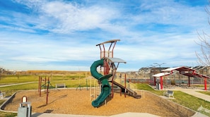 Une aire de jeux avec un toboggan et des structures d’escalade, entourée d’un parc avec des bancs et un bâtiment en arrière-plan.