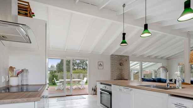 Une cuisine moderne avec des meubles blancs, un plan de travail en bois et un grand îlot central. On y trouve des suspensions vertes, une cheminée en briques et une vue sur la terrasse extérieure, avec une table et des chaises.