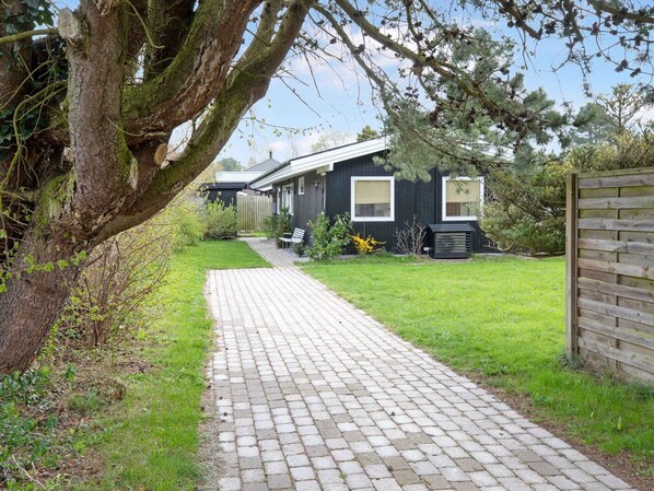 A pathway leading to a house with a dark exterior, a wooden fence, and a tree with hanging branches. - 5 Star Holiday Home in Hornbaek (Hornbaek)