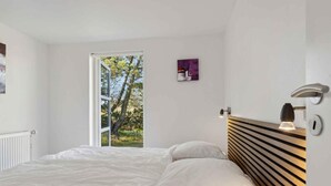 A bedroom with a large bed, a wooden headboard, a bedside table with a lamp, and a view of trees through an open door. - 5 Star Holiday Home in Saeby (Ronde)