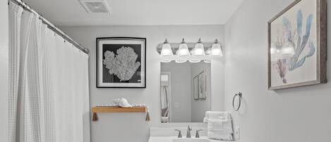 Un baño con inodoro blanco, un lavamanos con espejo y una cortina de ducha. Hay cuadros enmarcados en la pared y un perchero para toallas.