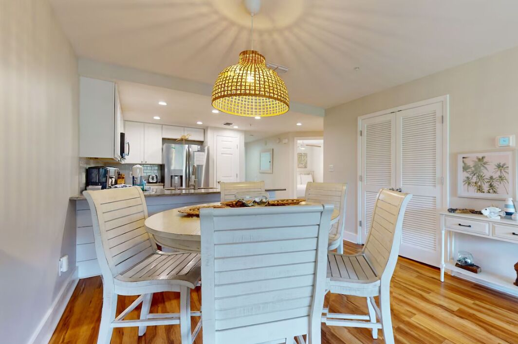 A dining area with a round table and four chairs, a chandelier, and a kitchen in the background.