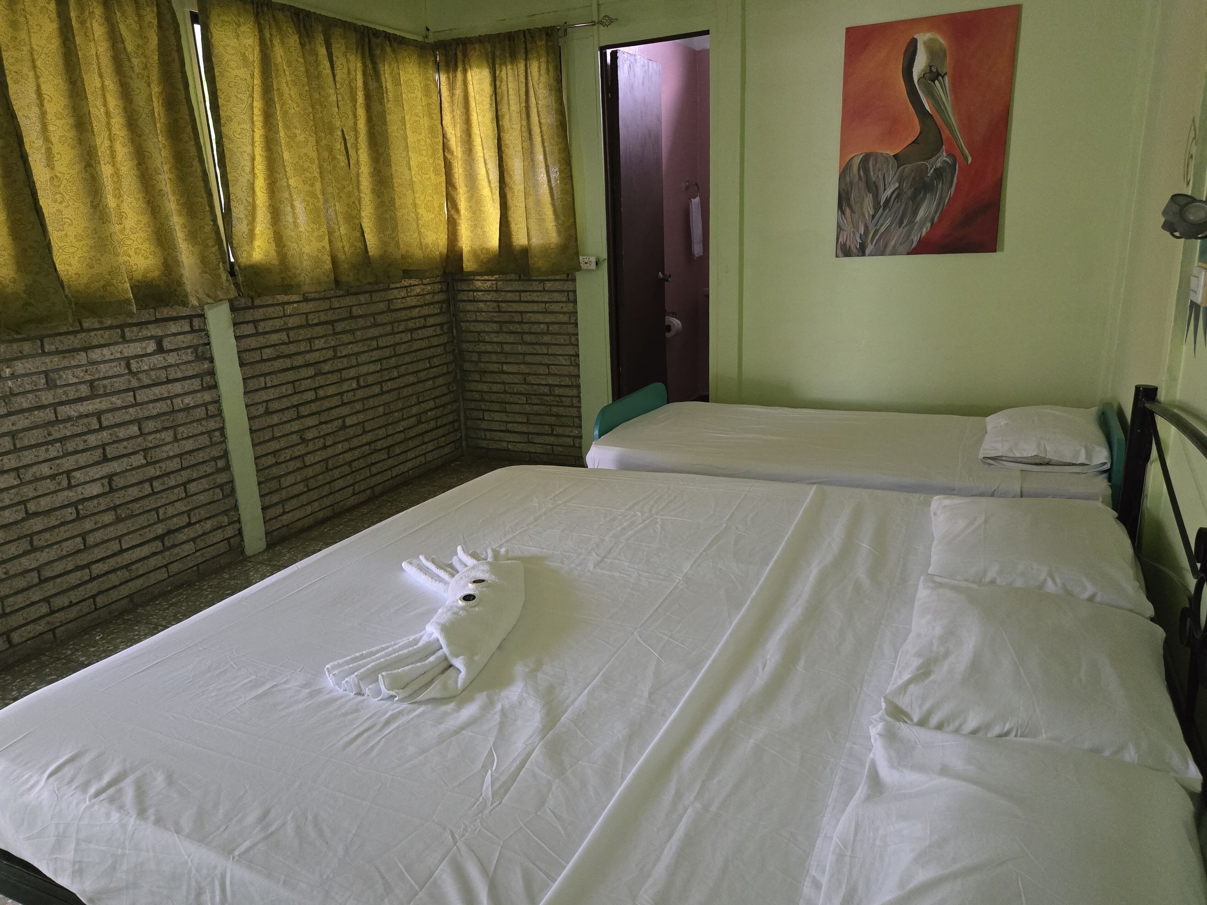 A hotel room with two beds, a brick wall, a painting, and a window with curtains.