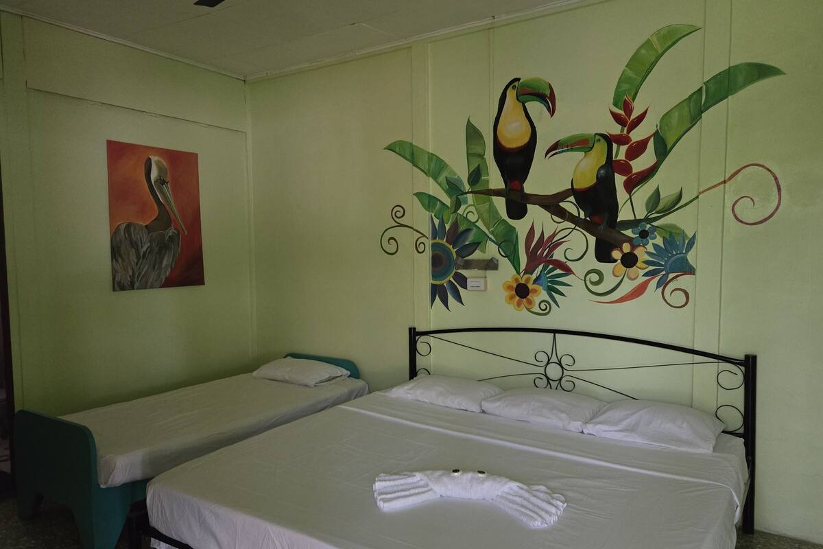 A hotel room with two single beds, a mural of toucans and flowers on the wall, and a painting of a bird.