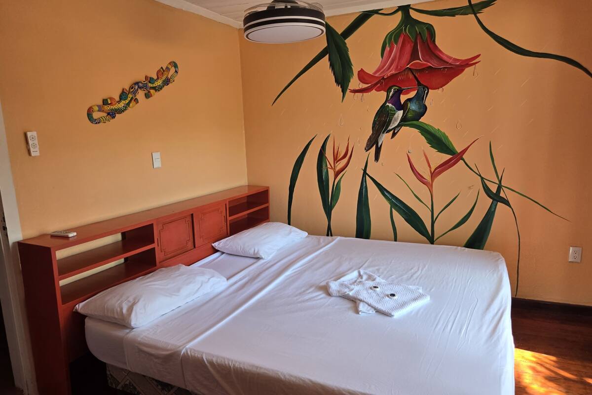 A hotel room with a bed, a wooden headboard, a wall mural, and a ceiling light.