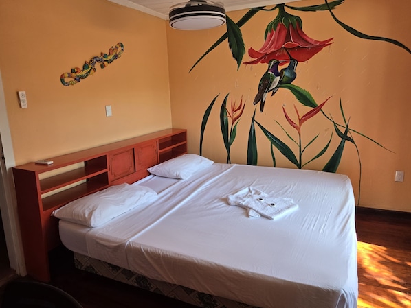 A hotel room with a bed, a wooden headboard, a wall mural, and a ceiling light. - Jane's Beach Resort (El Roble)