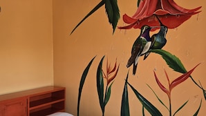 A hotel room with a bed, a wooden nightstand, and a mural of birds and flowers on the wall. - Jane's Beach Resort (El Roble)