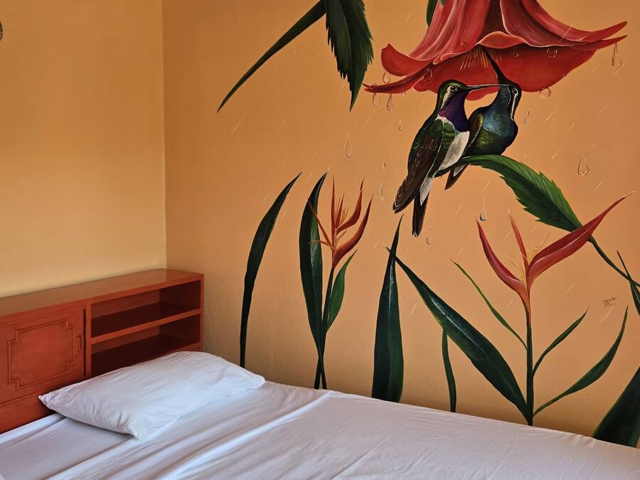 Habitación de hotel con cama, mesita de noche de madera y un mural de pájaros y flores en la pared.