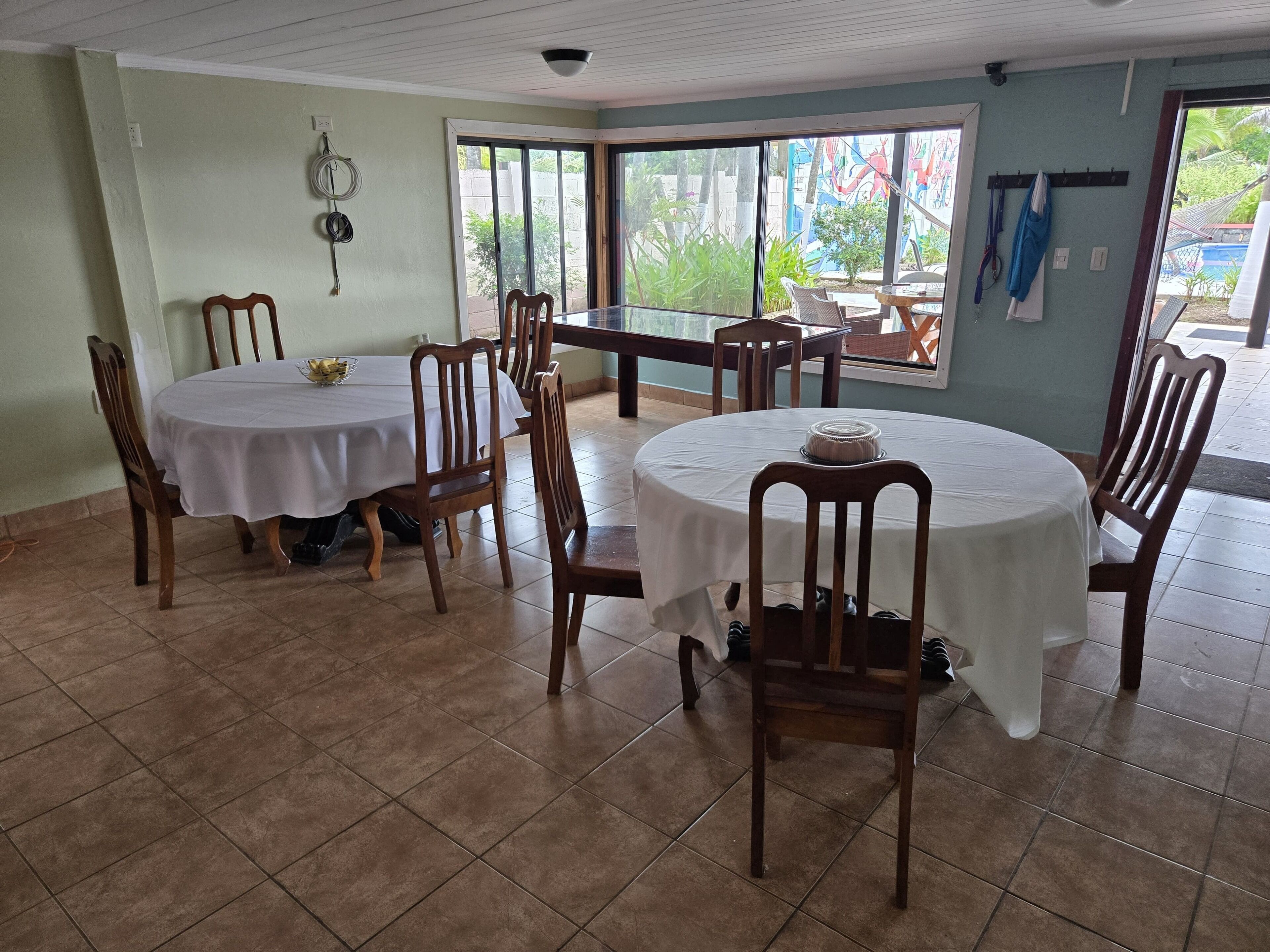 Comedor con mesas redondas y sillas de madera, suelo de baldosas y ventanas amplias con vista a la vegetación exterior.