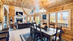 A cozy cabin interior with a fireplace, wooden walls, a dining area, and large windows.