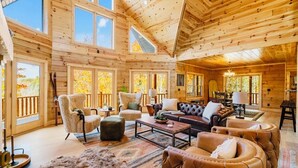 A spacious log cabin interior with multiple seating areas, large windows, and a high ceiling.