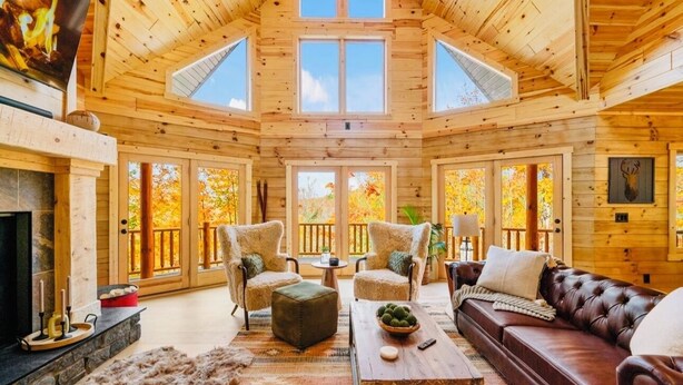 A log cabin interior with a fireplace, leather sofa, and multiple windows offering views of autumn foliage.