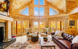 A log cabin interior with a fireplace, leather sofa, and multiple windows offering views of autumn foliage. - Sleeps 22. Minutes to Skiing at Nubs Nob & Highlands & Golf! Spacious & Private (Harbor Springs)
