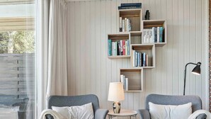 Ein gemütliches Zimmer mit zwei grauen Sesseln, einem kleinen runden Tisch mit Lampe und einem Bücherregal voller Bücher.