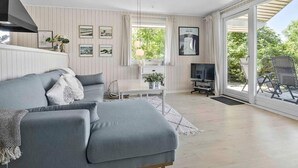 Ein modernes Wohnzimmer mit einem grauen Sofa, einem kleinen Tisch und einem Fernseher. An der Wand hängen Bilderrahmen und ein Fenster mit Vorhängen.