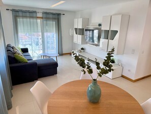 A modern living room with a sofa, a dining table with a vase, and a kitchen area with white cabinets and a TV. - Apartment Flor de Amendoeira Garden | 6min from the beach, parking and pool (Albufeira)