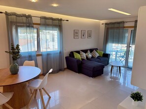 A modern living room with a sofa, a dining table, and chairs. There are curtains on the windows, and a balcony with outdoor furniture is visible. - Apartment Flor de Amendoeira Garden | 6min from the beach, parking and pool (Albufeira)