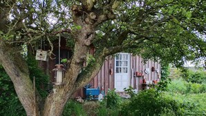 A rustic wooden house with a large tree in front, a white door, and various outdoor items like a lantern and pots. - 4 Star Holiday Home in Kirke Hyllinge (Kirke Hyllinge)