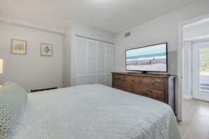 A bedroom with a bed, a dresser, a television displaying a beach scene, a closet, and a door leading to a balcony.