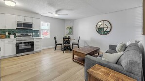 Eine moderne Küche mit weißen Schränken, einer Mikrowelle und einem Backofen. Es gibt eine Essecke mit Tisch und Stühlen sowie ein graues Sofa im Wohnzimmer.
