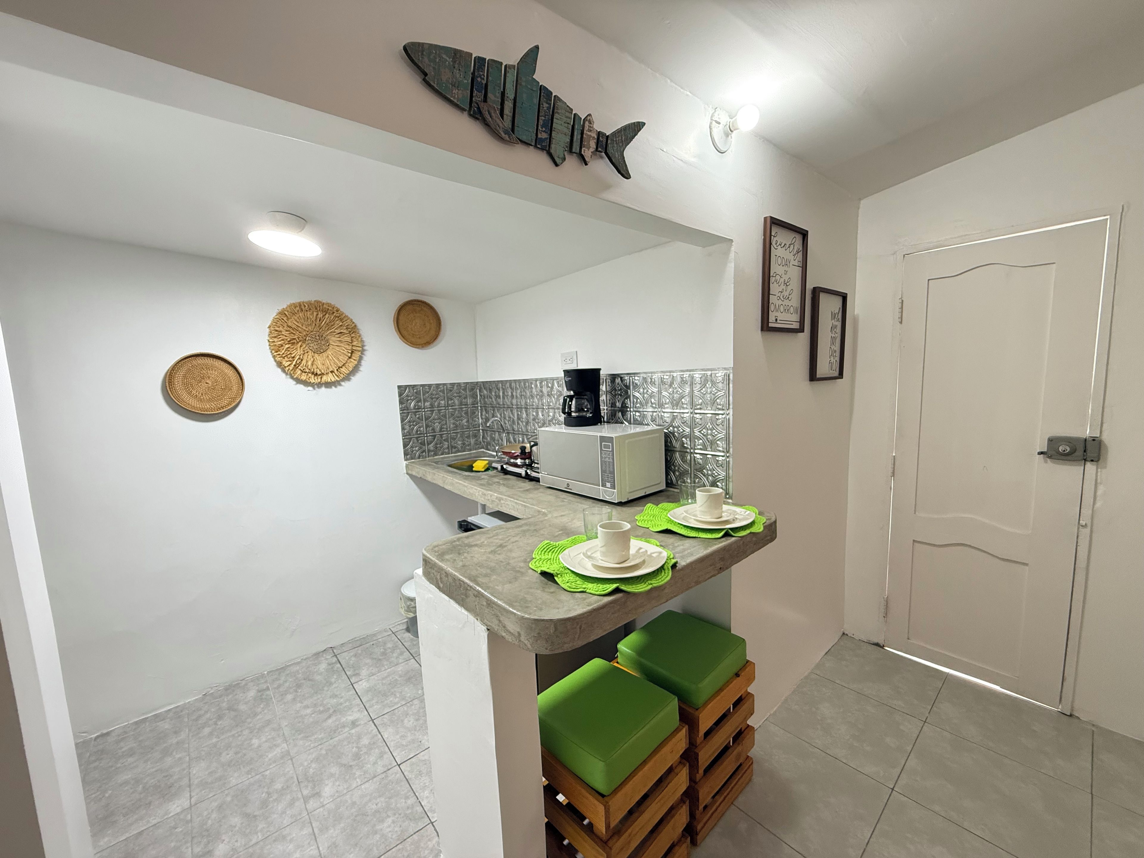 Cocina compacta con microondas, cafetera y un lavamanos pequeño. Se observan adornos en la pared y una cita enmarcada.