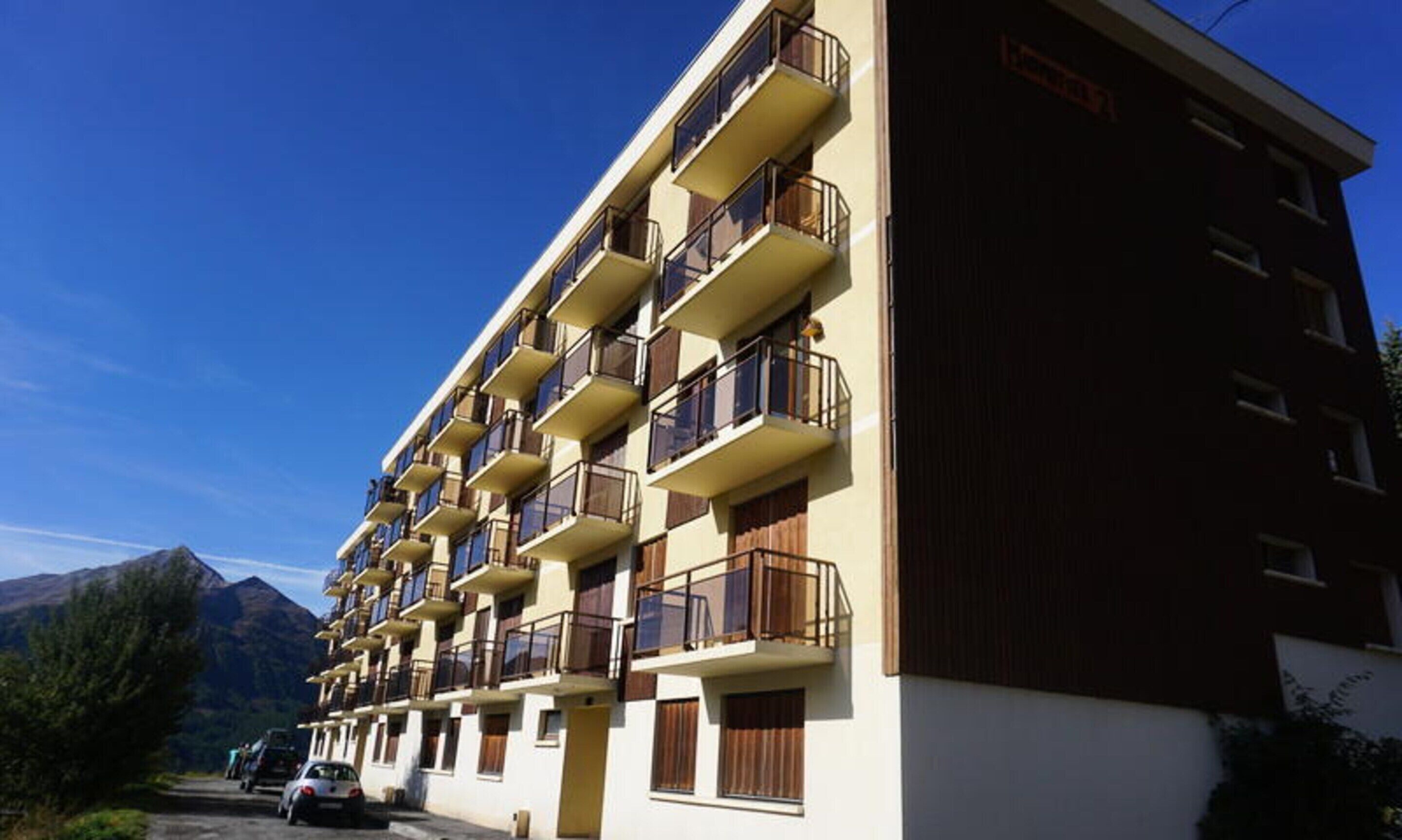 A multi-story hotel with balconies, a mountain backdrop, and a parking area.
