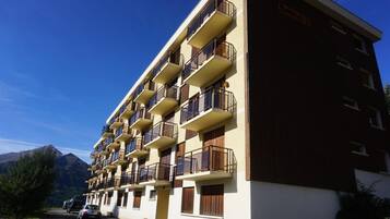 A multi-story hotel with balconies, a mountain backdrop, and a parking area.