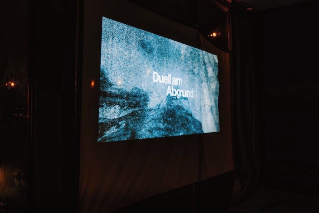 A projection screen displaying the text "Duell am Abgrund" in a dark room.