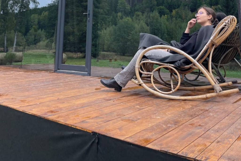 A person sitting on a wicker rocking chair on a wooden deck with a glass door leading to a forested area.