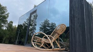 Een modern gebouw met een glazen gevel die reflecteert, een houten terras en een rieten stoel.