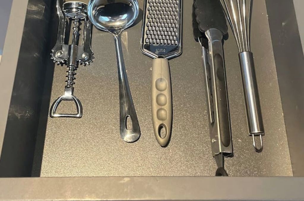A drawer containing various kitchen utensils including a grater, a spoon, a ladle, a bottle opener, and a pair of tongs.