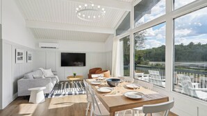 A modern living room with a wooden dining table set for six, a grey sofa, a flat-screen TV mounted on the wall, and a large window offering a view of a lake and trees. - Lakelyn Lodge (Lakeville)