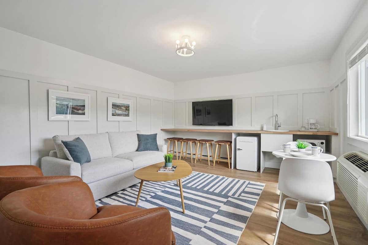 A modern living room with a sofa, coffee table, and chairs, a kitchen area with a dining table, and a TV mounted on the wall.