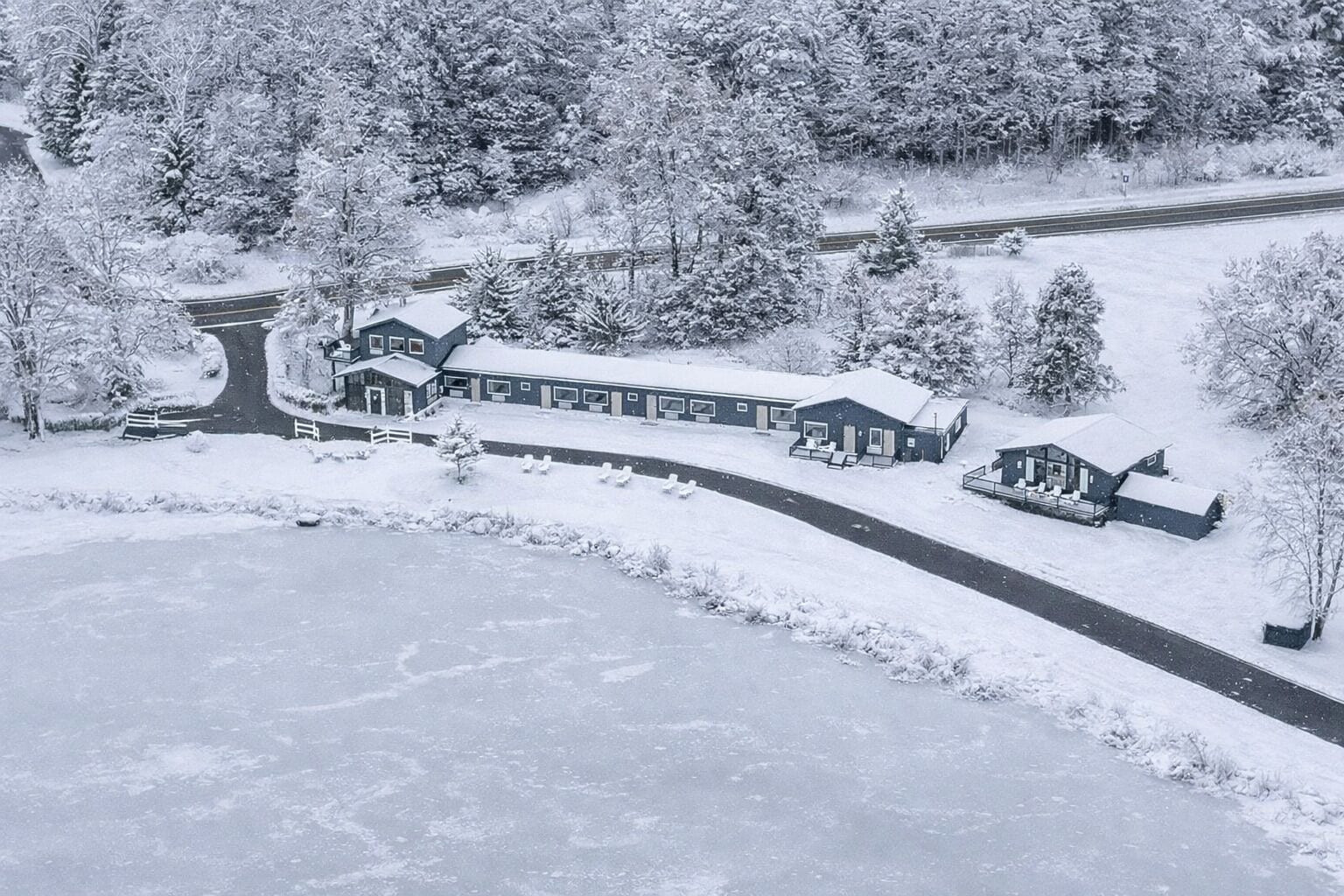 Un grand chalet enneigé, avec une longue allée menant à l’entrée, entouré d’une forêt dense.