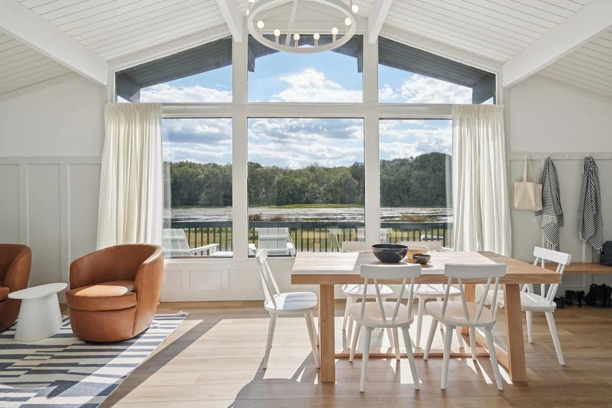 A dining area with a large table, white chairs, and a view of a forest through large windows.