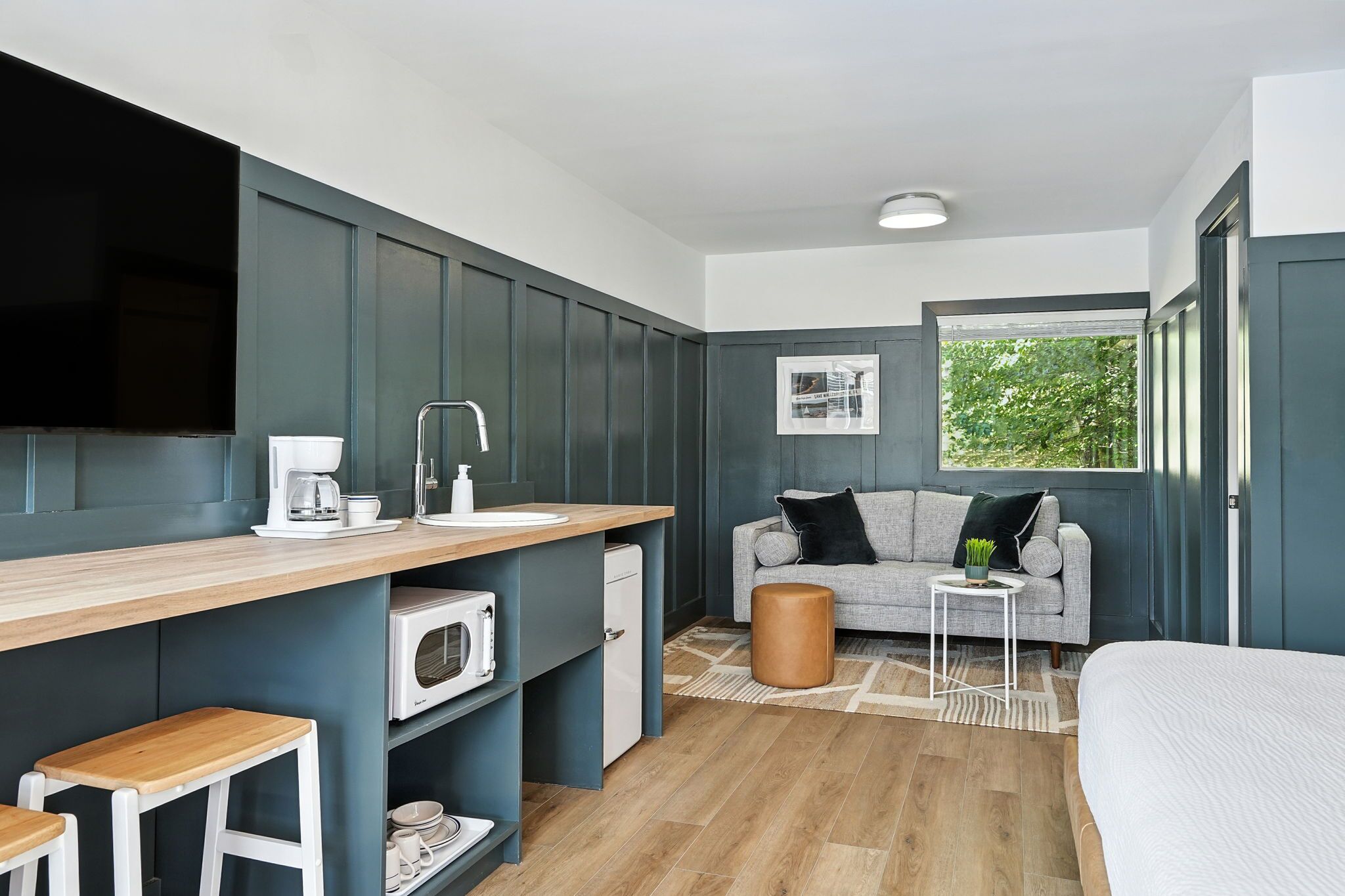 A compact room with a kitchenette, a sofa, a bed, and a window with a view of greenery.