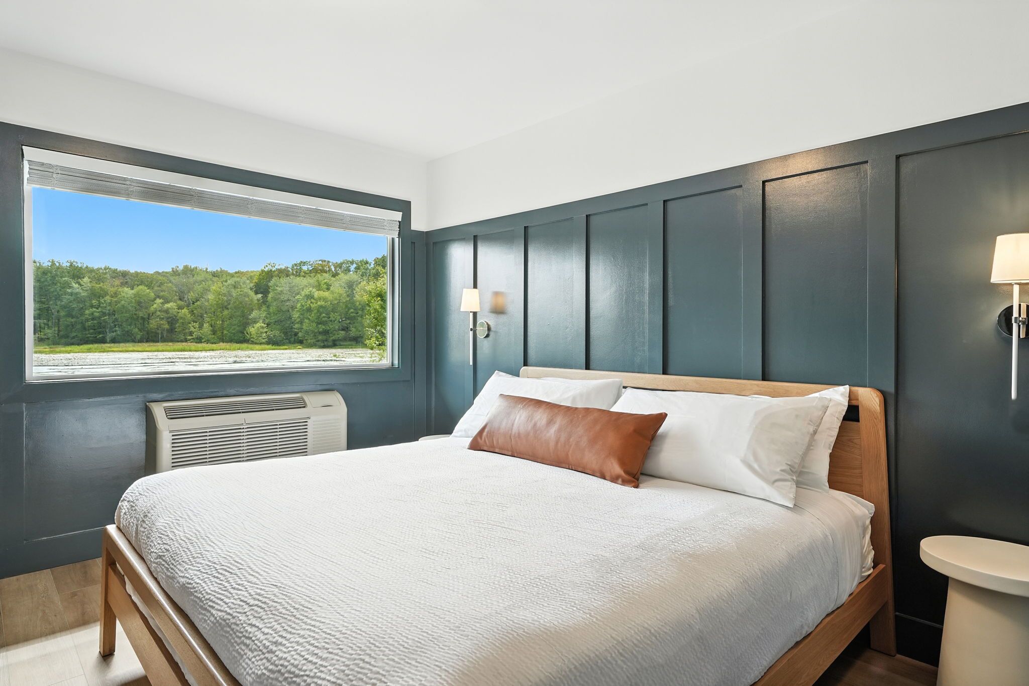 A bedroom with a large window, a bed with white and brown bedding, and a wall-mounted lamp.