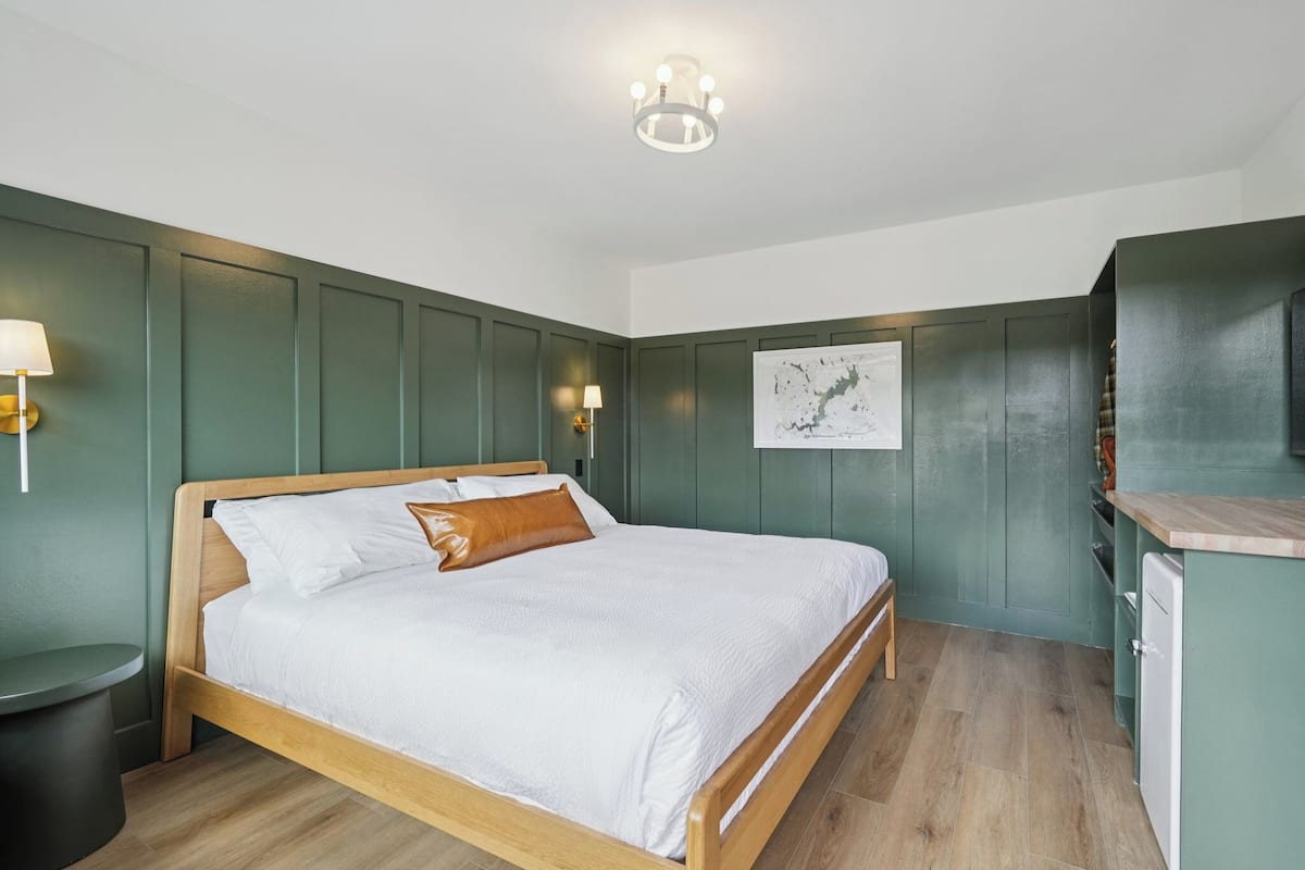 A bedroom with a wooden bed, a small bedside table, a wall-mounted lamp, and a green-painted wardrobe.