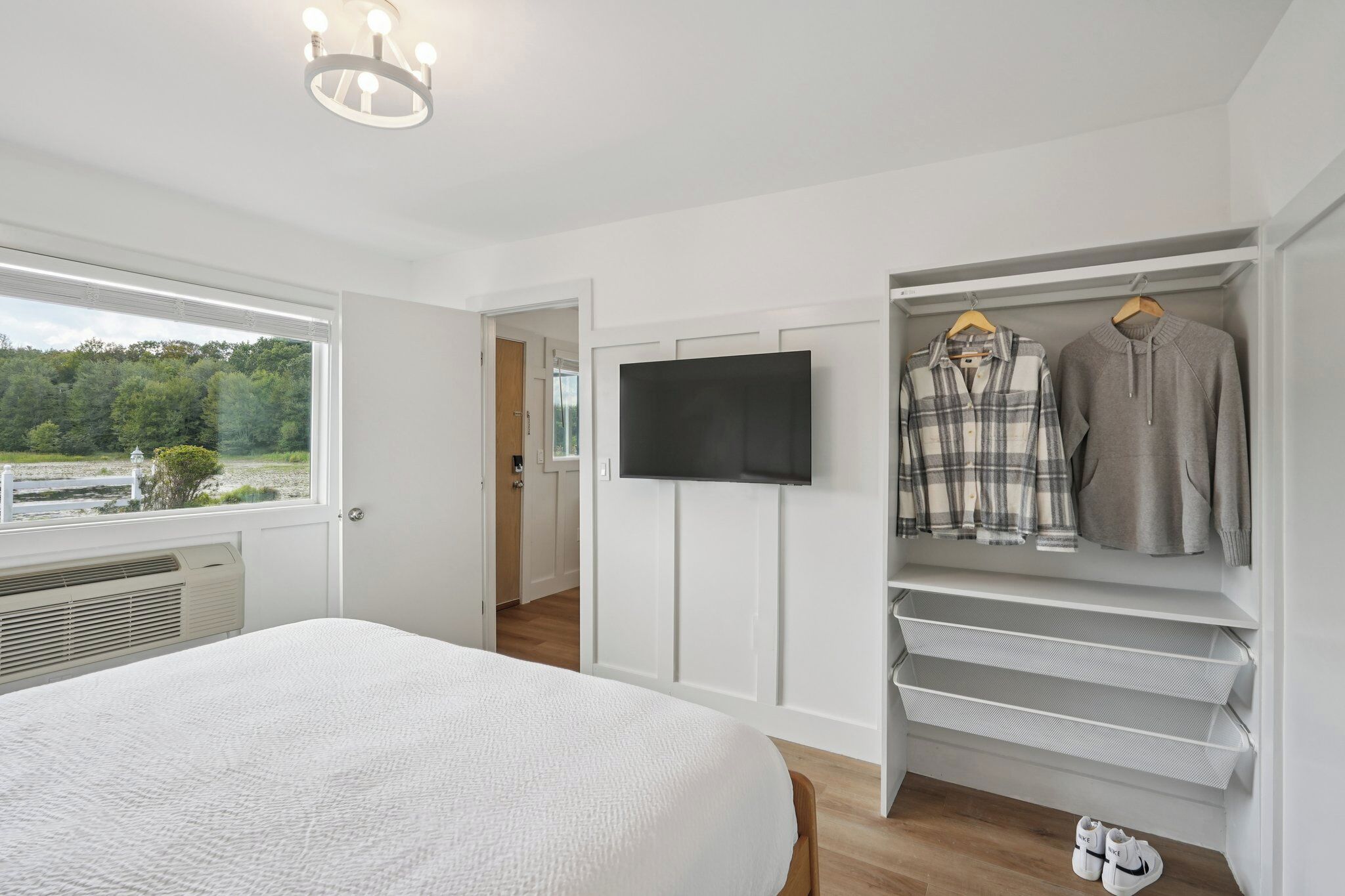 Un dormitorio con cama, televisor montado en la pared, armario con ropa colgada y ventana con vistas a árboles.