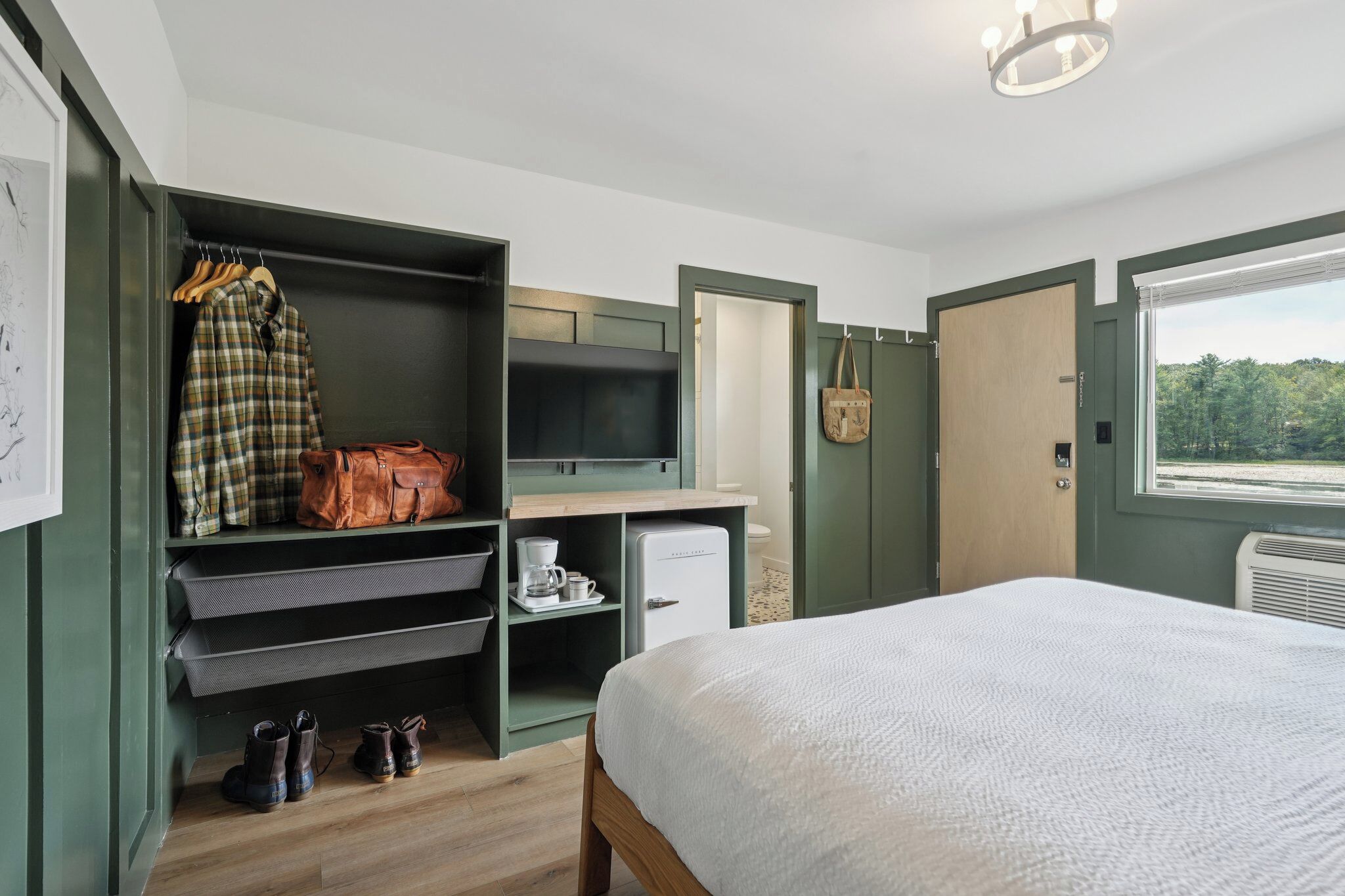 A bedroom with a bed, a wardrobe, a small kitchenette, and a window with a view of trees.