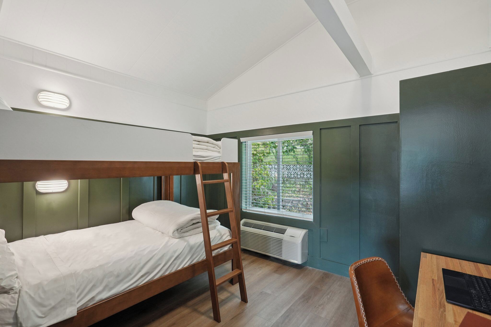 A bunk bed with a ladder, a desk with a chair, a window with a view of greenery, and a ceiling with recessed lighting.
